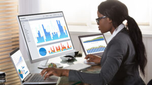 a working professional sitting behind three monitors all showing graphical data in different chart types